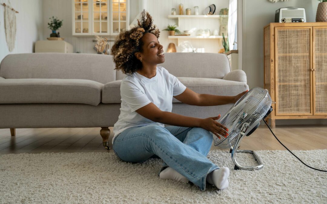 black woman sitting on the floor in front of a fan trying to cool off.
